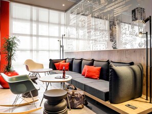 A modern lounge area with a sofa, chairs, a table, a plant, and a wall with artistic designs. - ibis Muenchen City (Munich)