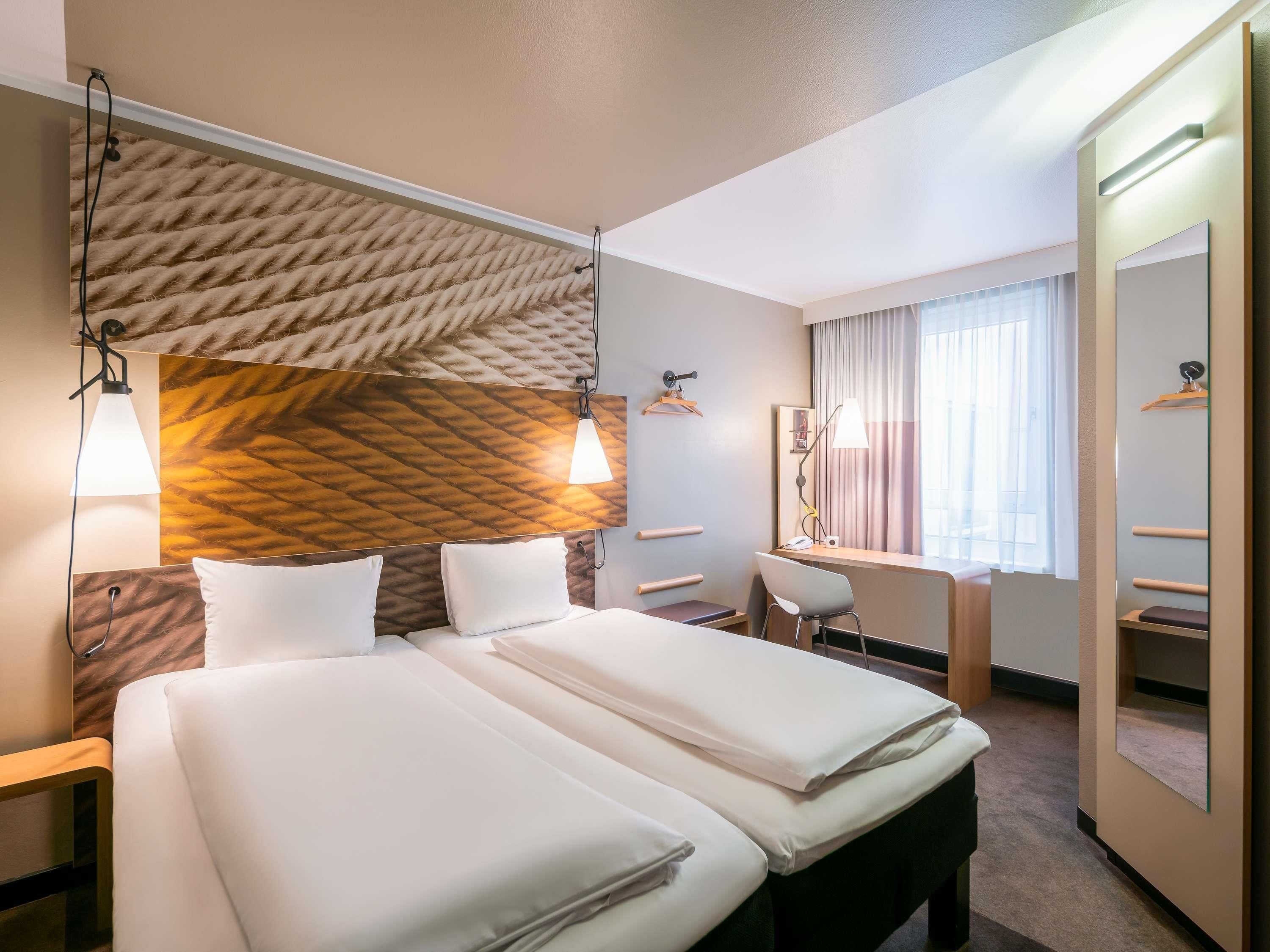 Habitación de hotel con dos camas, un escritorio, una silla, un espejo y una ventana con cortinas.