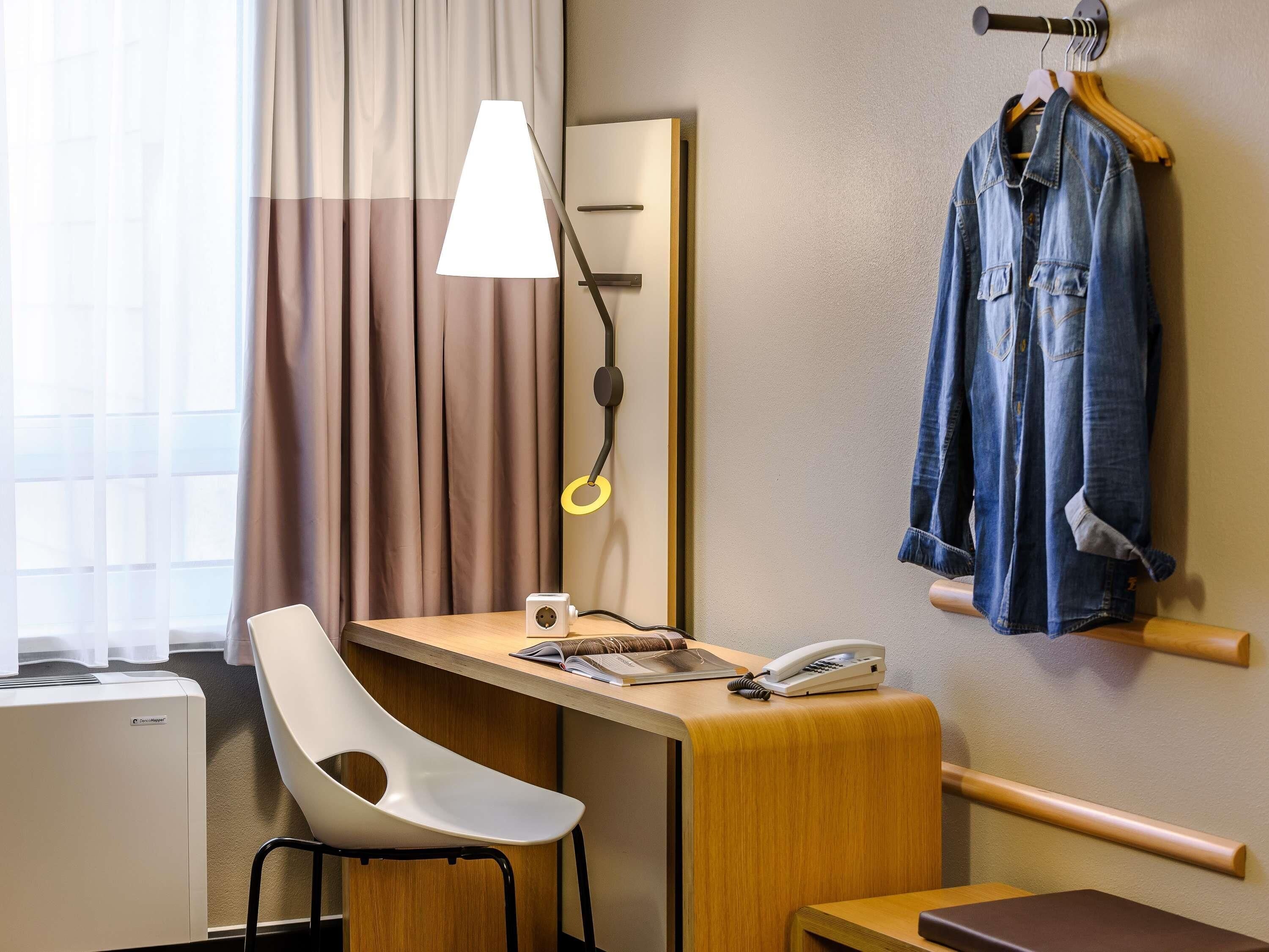 A hotel room with a desk, chair, lamp, phone, and a hanging jacket.