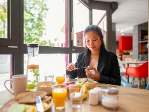 Una persona desayunando en una mesa, con objetos de desayuno, una ventana que da a zonas verdes y sillas rojas al fondo.
