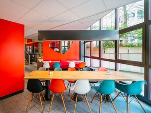 Un comedor moderno con una larga mesa de madera y sillas coloridas, ventanas amplias que ofrecen vistas al exterior y una pared de acento roja y vibrante.