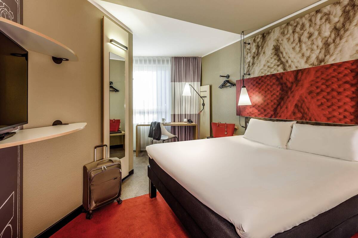 A hotel room with a bed, a desk, a chair, a TV, a suitcase, and a coat rack.