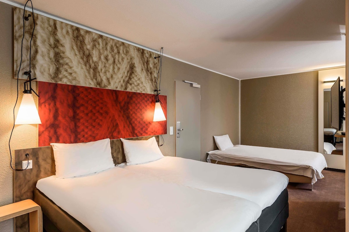 A hotel room with a large bed, two bedside tables, a mirror, and a red accent wall.