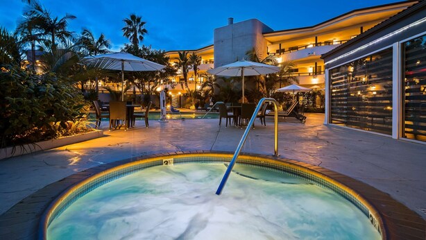 A heated outdoor pool area with lounge chairs and umbrellas, surrounded by palm trees and a building with multiple balconies.