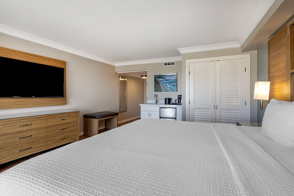 Quarto de hotel com uma cama grande, televisão fixada na parede, escrivaninha com cadeira, um quadro na parede e um guarda-roupa.