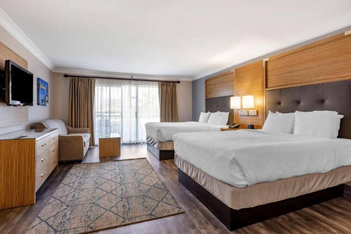 Quarto de hotel com duas camas, uma escrivaninha, televisão e uma janela grande com cortinas.