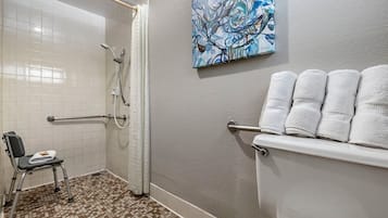 A bathroom with a shower, a toilet, and a chair. There is a towel rack with towels and a painting on the wall.
