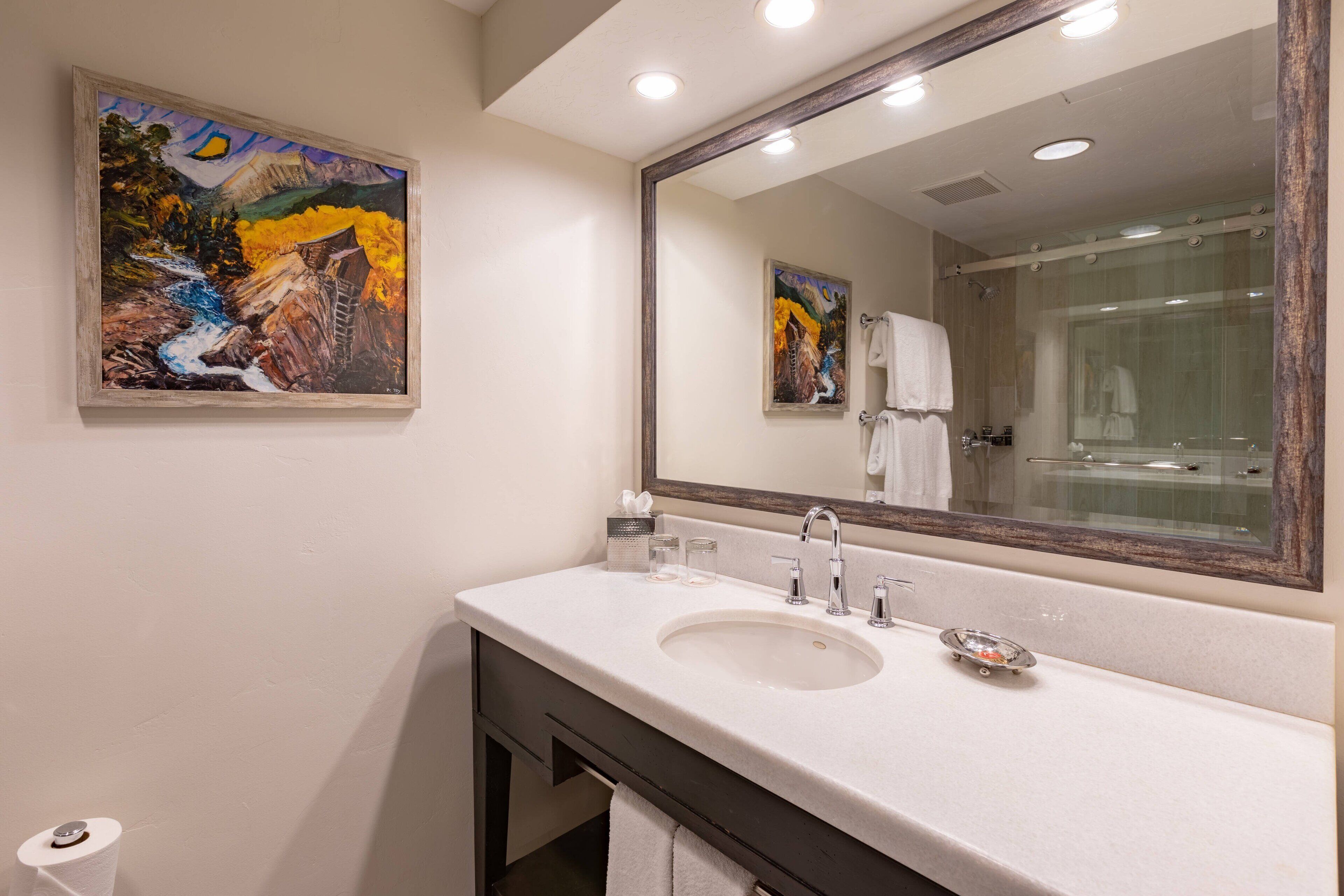 A bathroom with a large mirror, a sink, and a painting of a landscape.