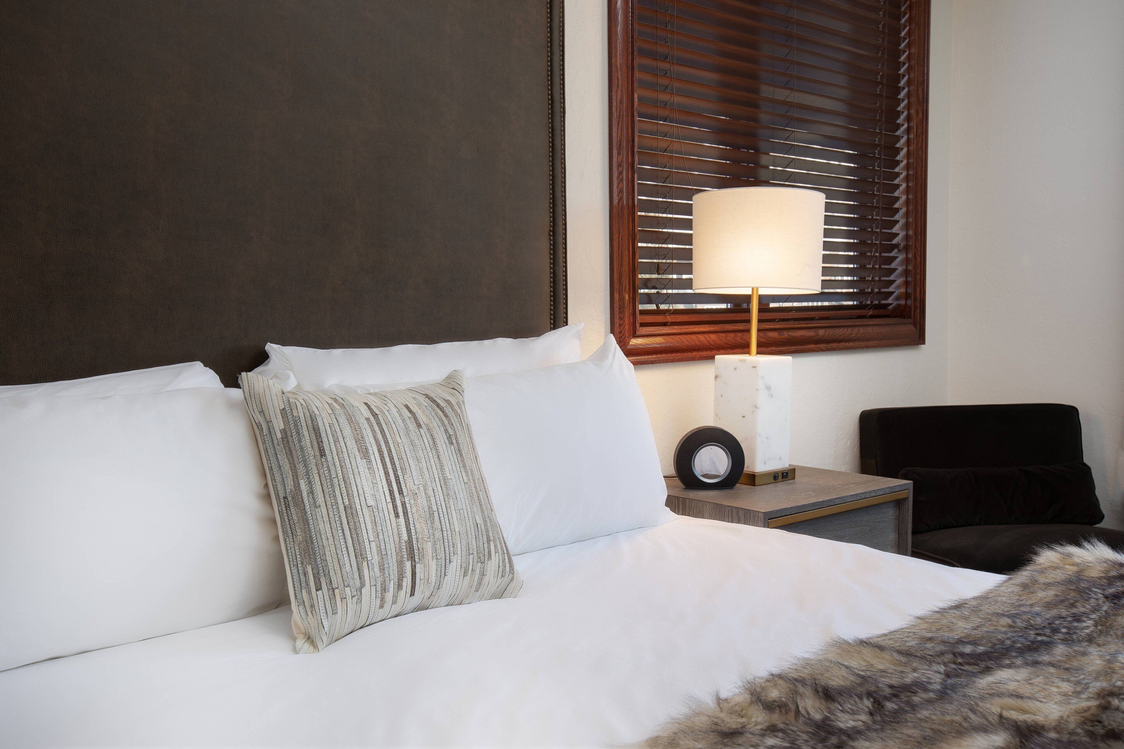 Uma cama bem arrumada com roupa de cama branca, um travesseiro texturizado, uma cabeceira de madeira, uma mesa de cabeceira com um abajur e uma cadeira.