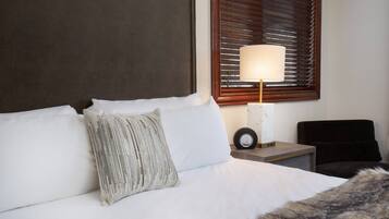 Una cama bien hecha con sábanas blancas, un cojín texturizado, un cabecero de madera, una mesita de noche con una lámpara y una silla.