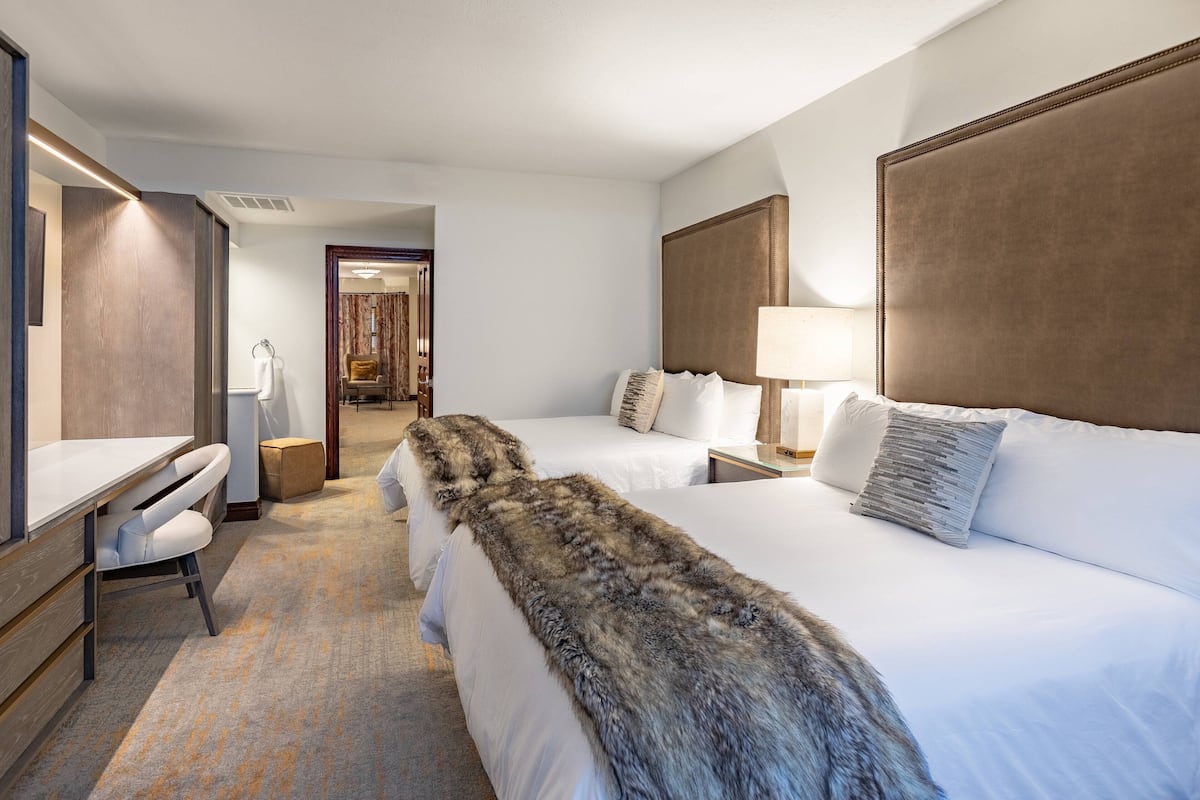 A hotel room with two beds, a desk, a chair, a fur throw, and a lamp.