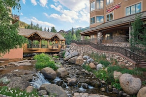 A lodge with a stone facade, a wooden bridge over a stream, and a mountain backdrop. - Beaver Creek Lodge, Autograph Collection (Avon)