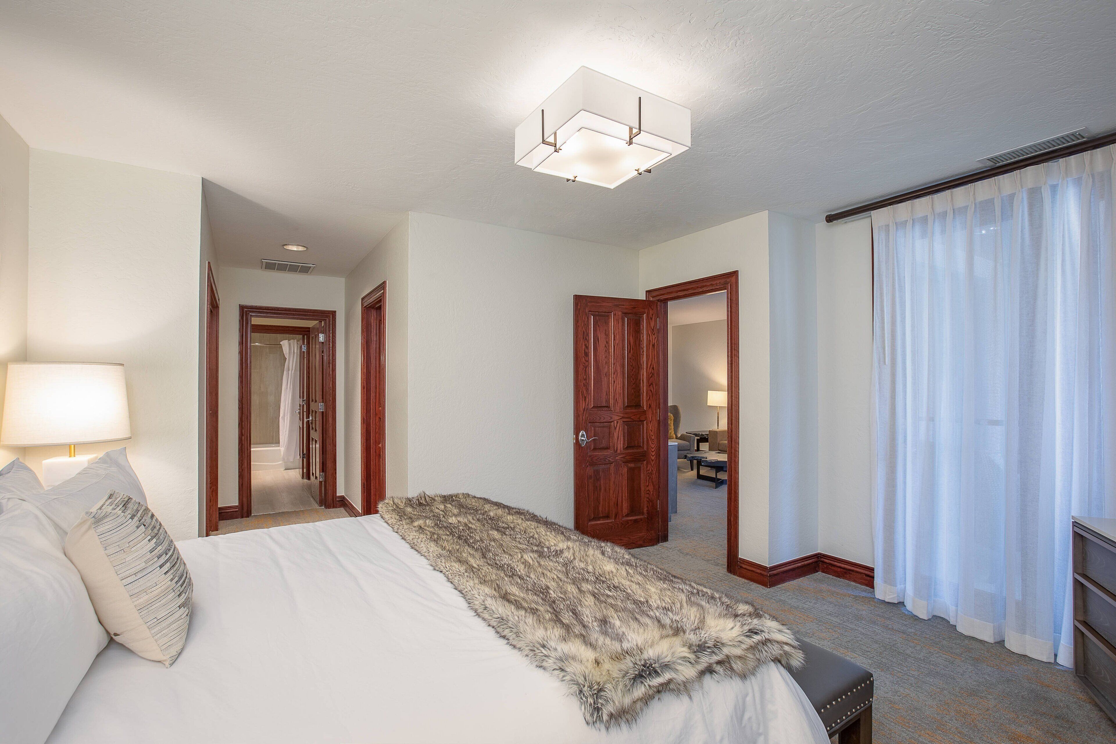 A hotel room with a large bed, a fur throw blanket, a wooden door, and a view of the hallway.