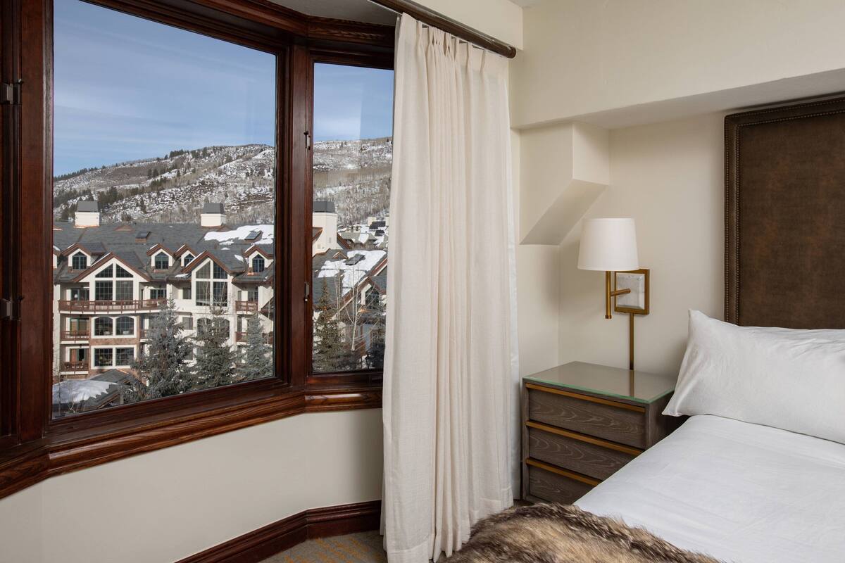 A hotel room with a large window, a bed, a bedside table with a lamp, and a view of a snowy landscape.