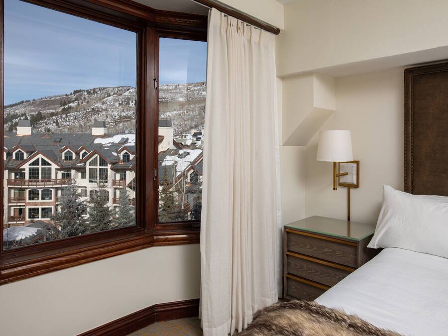A hotel room with a large window, a bed, a bedside table with a lamp, and a view of a snowy landscape.