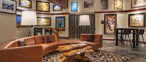 A spacious living room with a curved brown leather sofa, a wooden coffee table, and a checkered rug. The room features a high ceiling with exposed beams, a chandelier, and artwork on the walls.