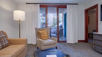 A hotel room with a sofa, armchair, and a coffee table with a magazine. There is a large window with curtains, a door leading to a balcony, and a dresser.