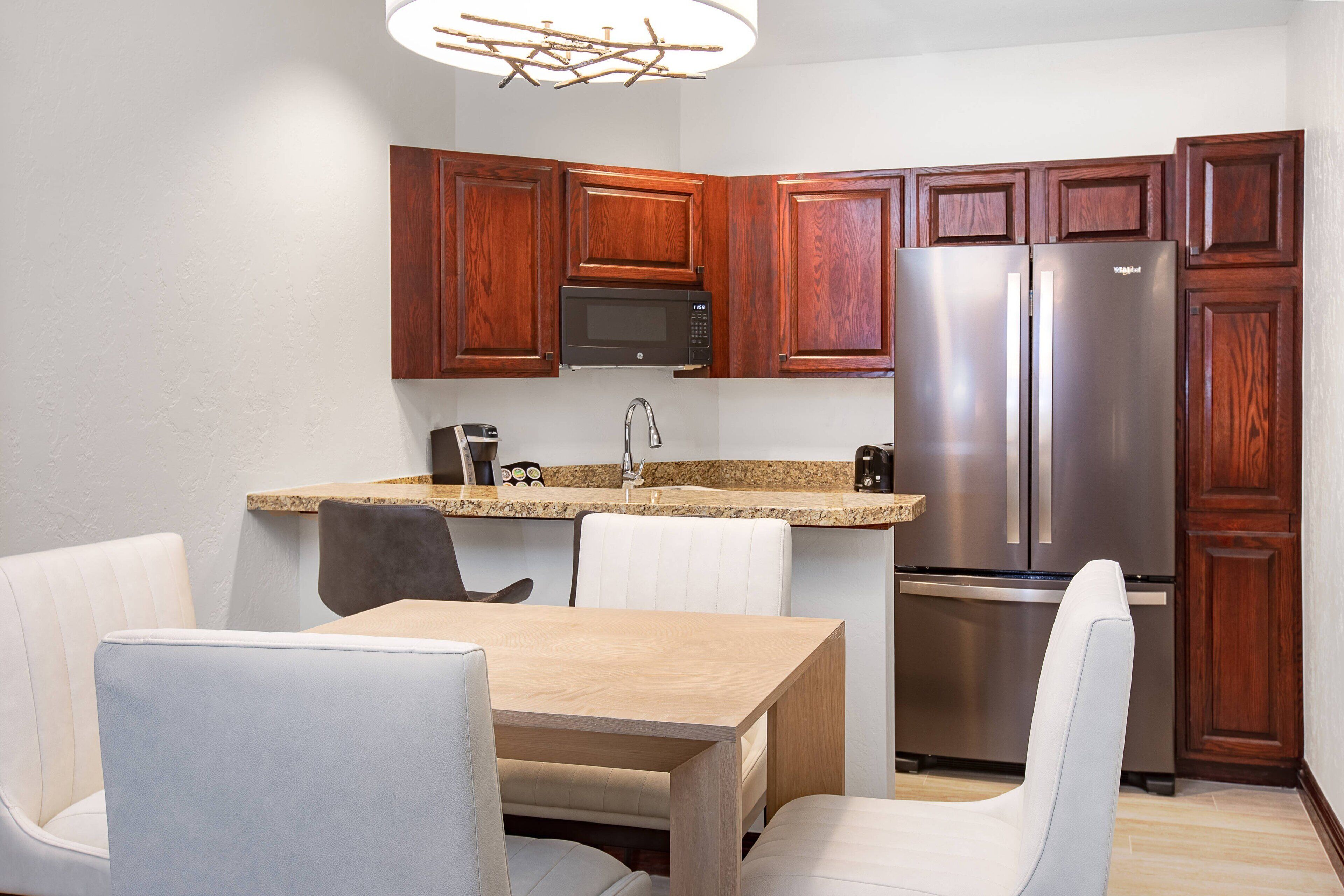 A modern kitchen with wooden cabinets, a stainless steel refrigerator, a microwave, and a dining area with a wooden table and white chairs.