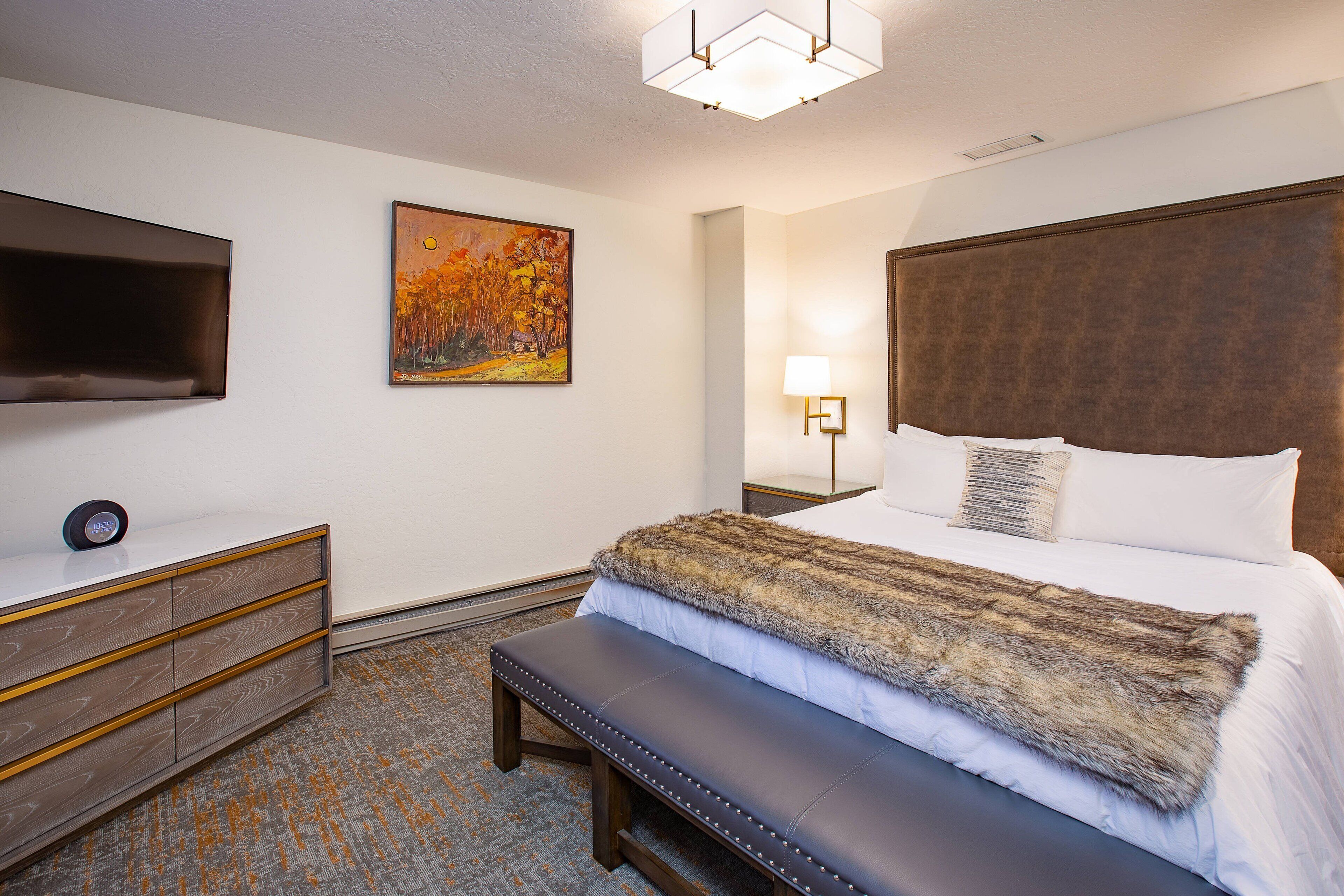 Habitación de hotel con una cama grande, un banco, un televisor, un cuadro y una mesita de noche.