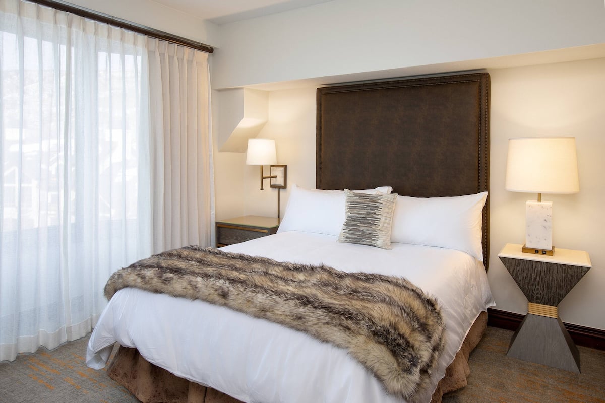 A hotel room with a large bed, a fur throw, two bedside lamps, and a view of the outdoors through sheer curtains.