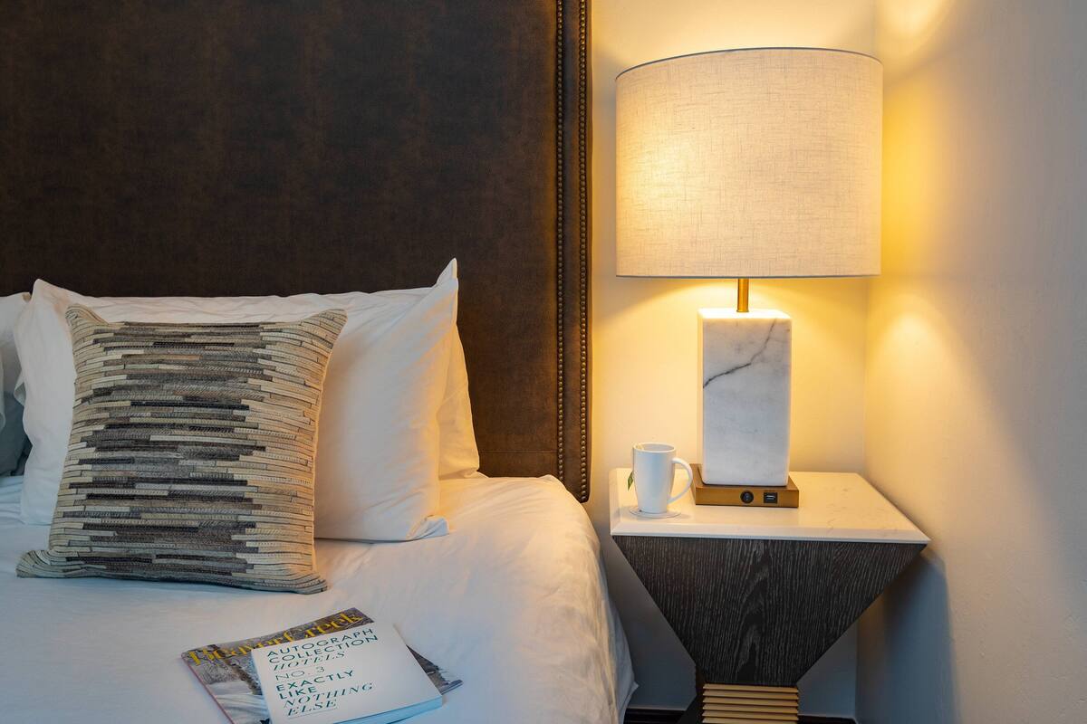 A neatly made bed with a patterned pillow, a bedside table with a lamp, and a magazine on the bed.