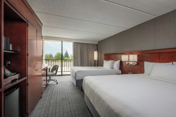 A hotel room with two beds, a desk, a chair, a minibar, and a view of the outdoors.