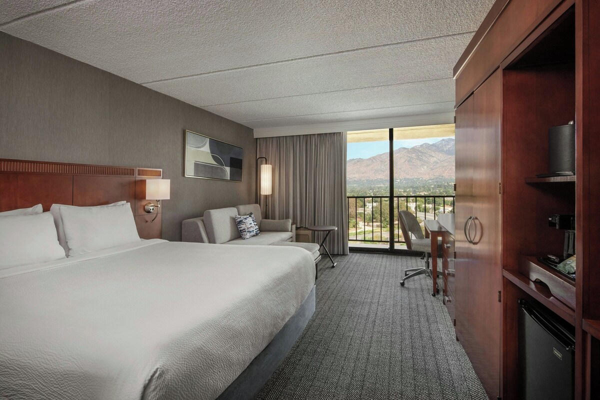 Une chambre d’hôtel avec un grand lit, un bureau, une chaise, une télévision et un balcon offrant une vue sur la montagne.