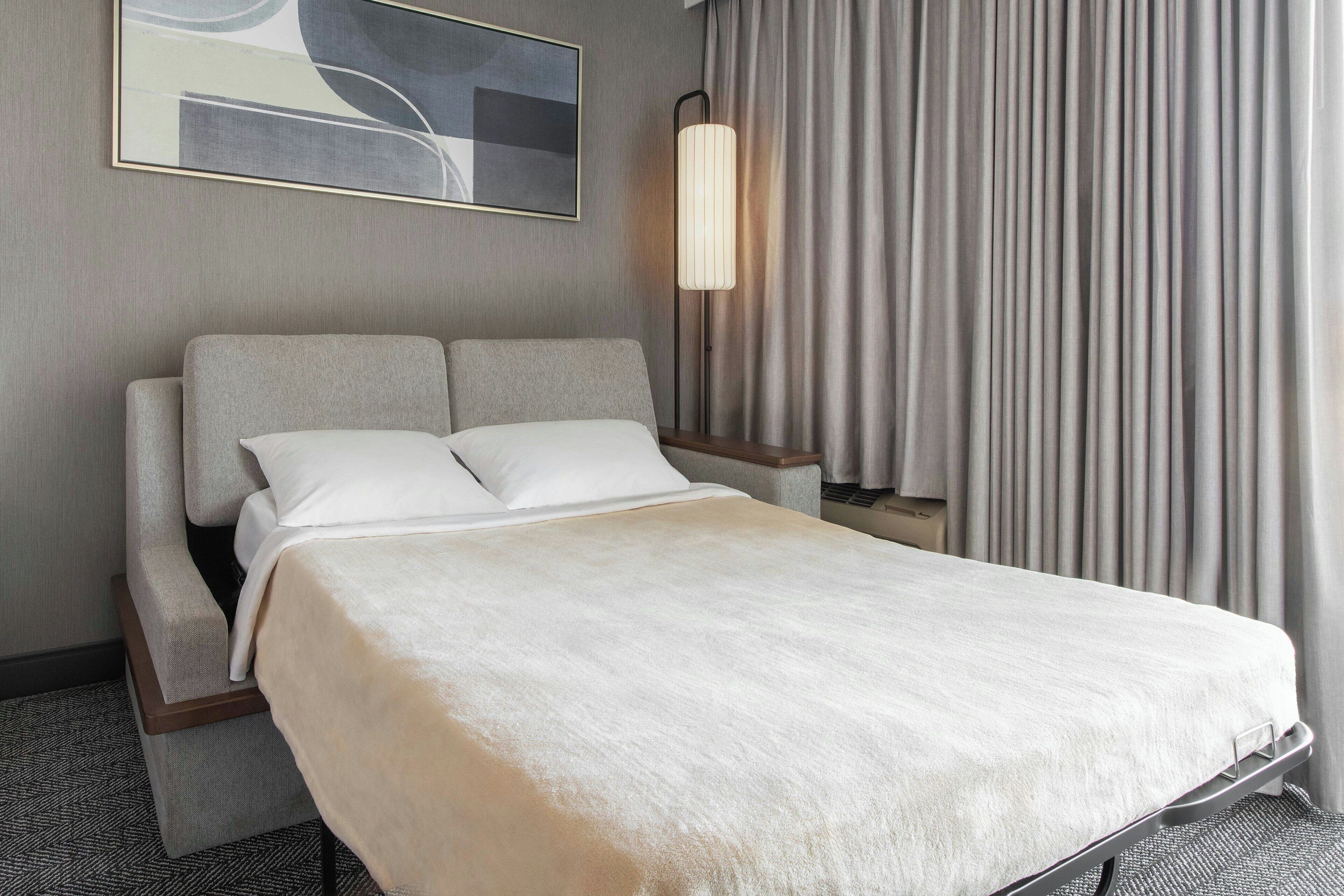 Habitació d'hotel amb llit, una taula de nit, una làmpada i una finestra amb cortines.