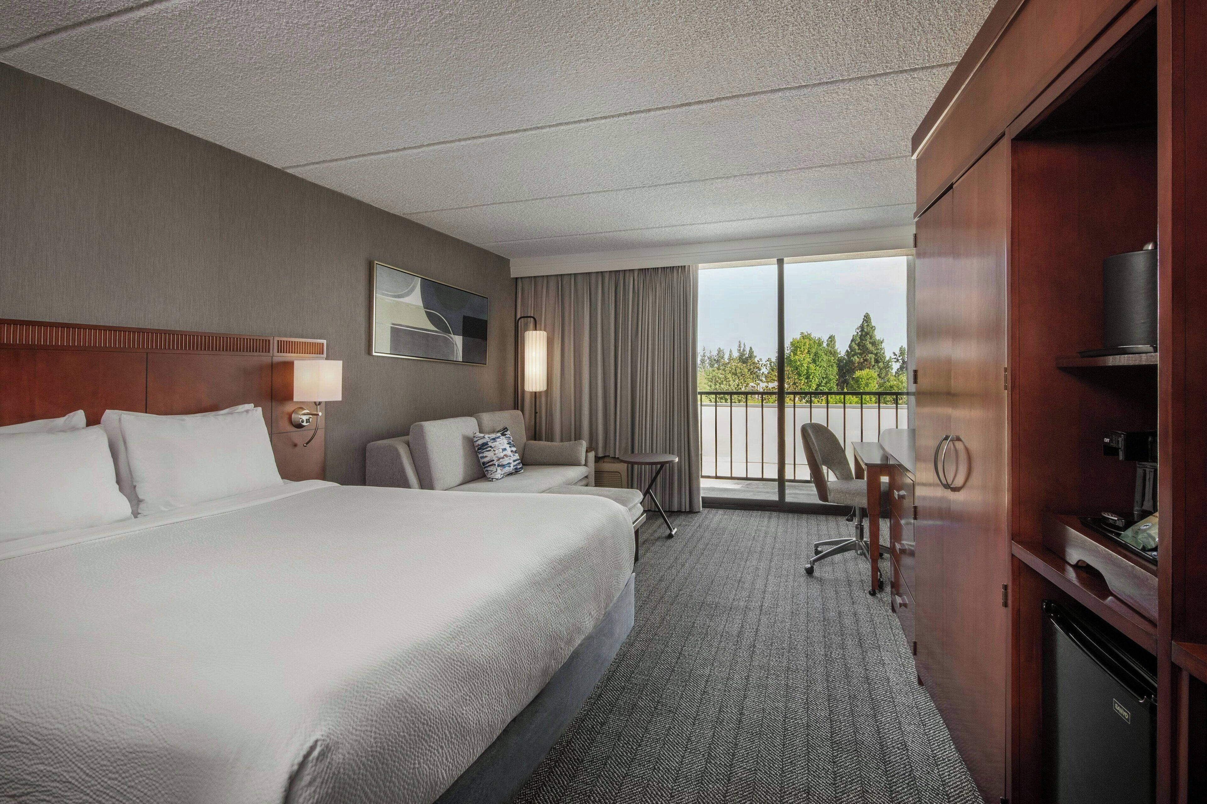 Habitación de hotel con una cama grande, un escritorio, una silla, un televisor y un balcón con vistas a árboles.