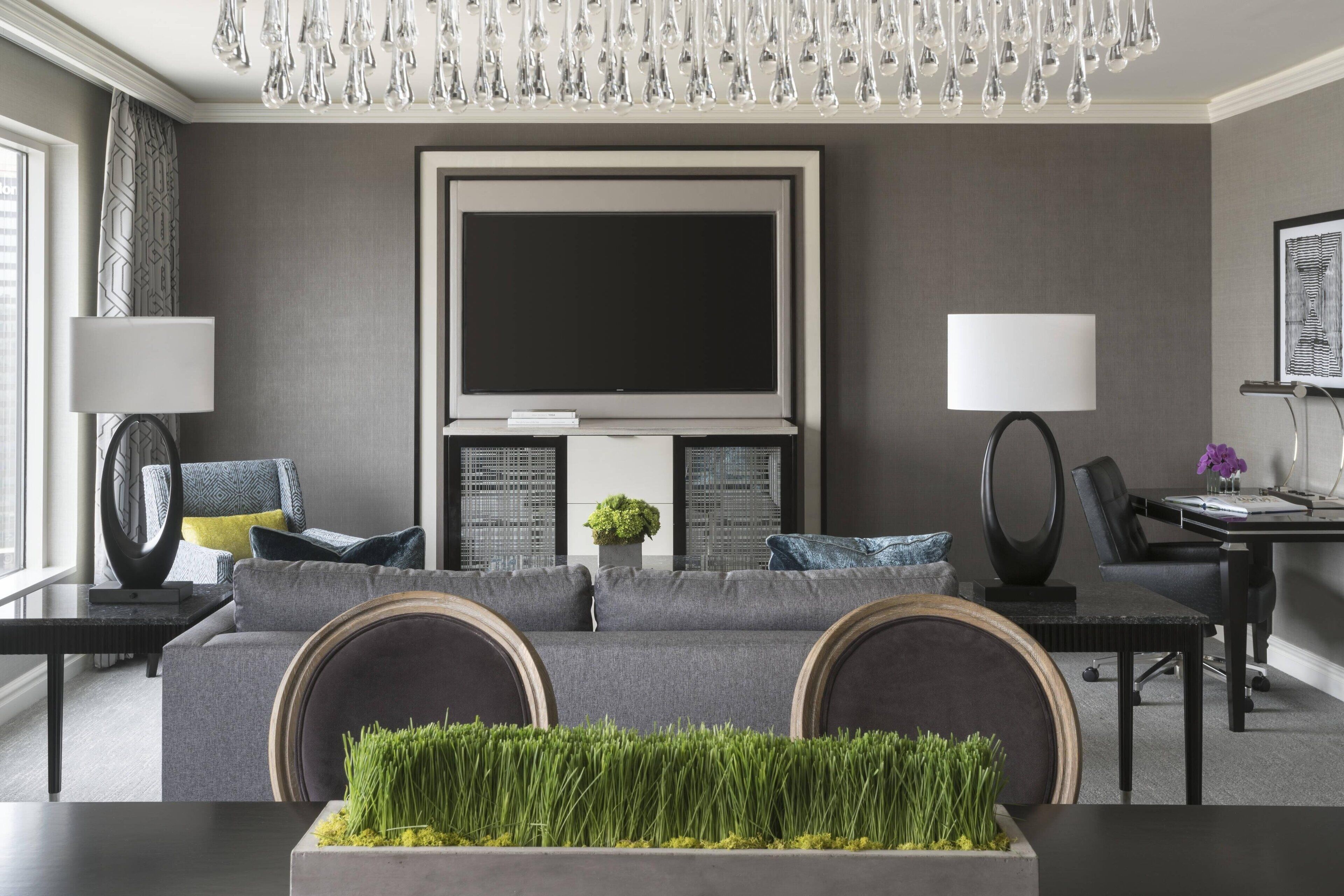 A modern living room with a grey sofa, a flat-screen TV, a dining table, and a chandelier.