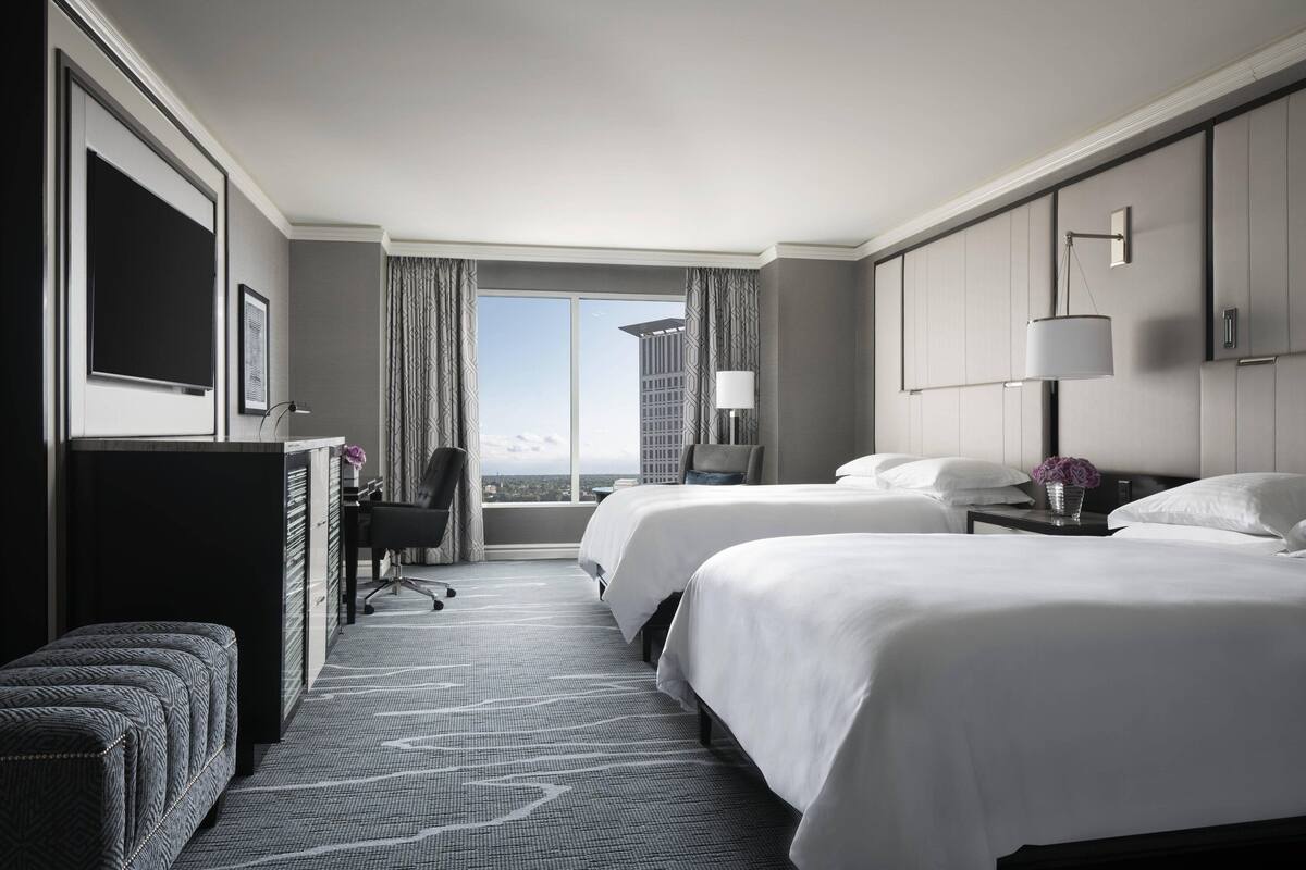 A hotel room with two beds, a desk, a chair, a TV, and a window with a city view.