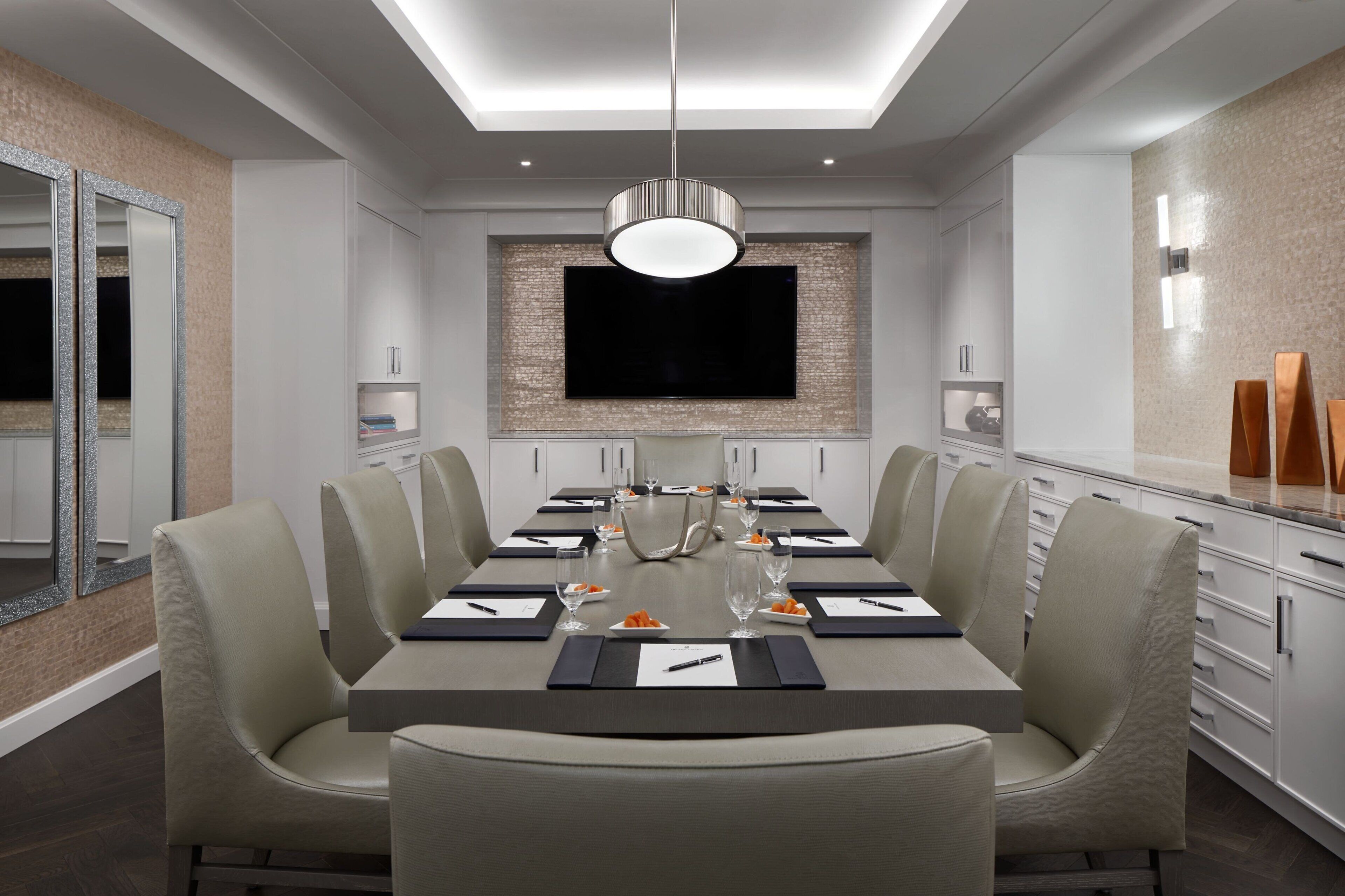 A modern dining room with a long table, comfortable chairs, a flat-screen TV mounted on the wall, and a pendant light above the table.