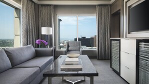 A modern hotel room with a sofa, a coffee table, a TV, and a view of the city.