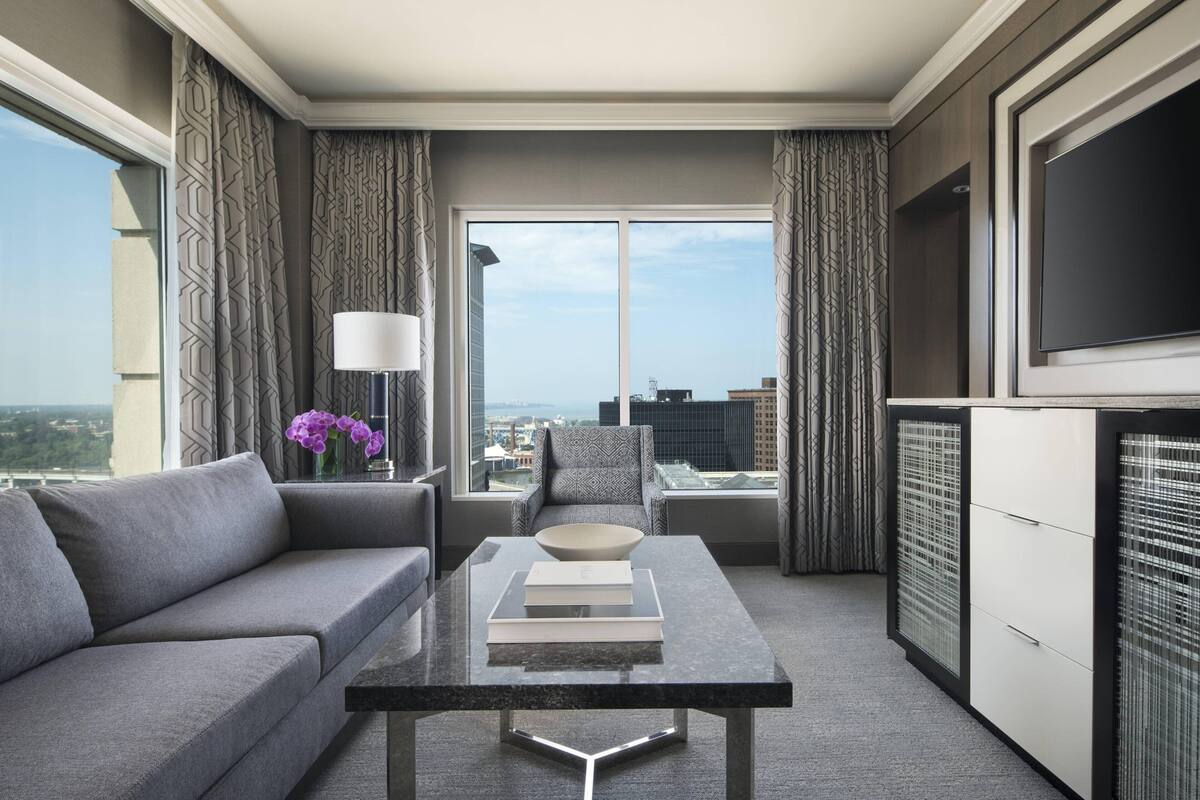 A modern hotel room with a sofa, a coffee table, a TV, and a view of the city.