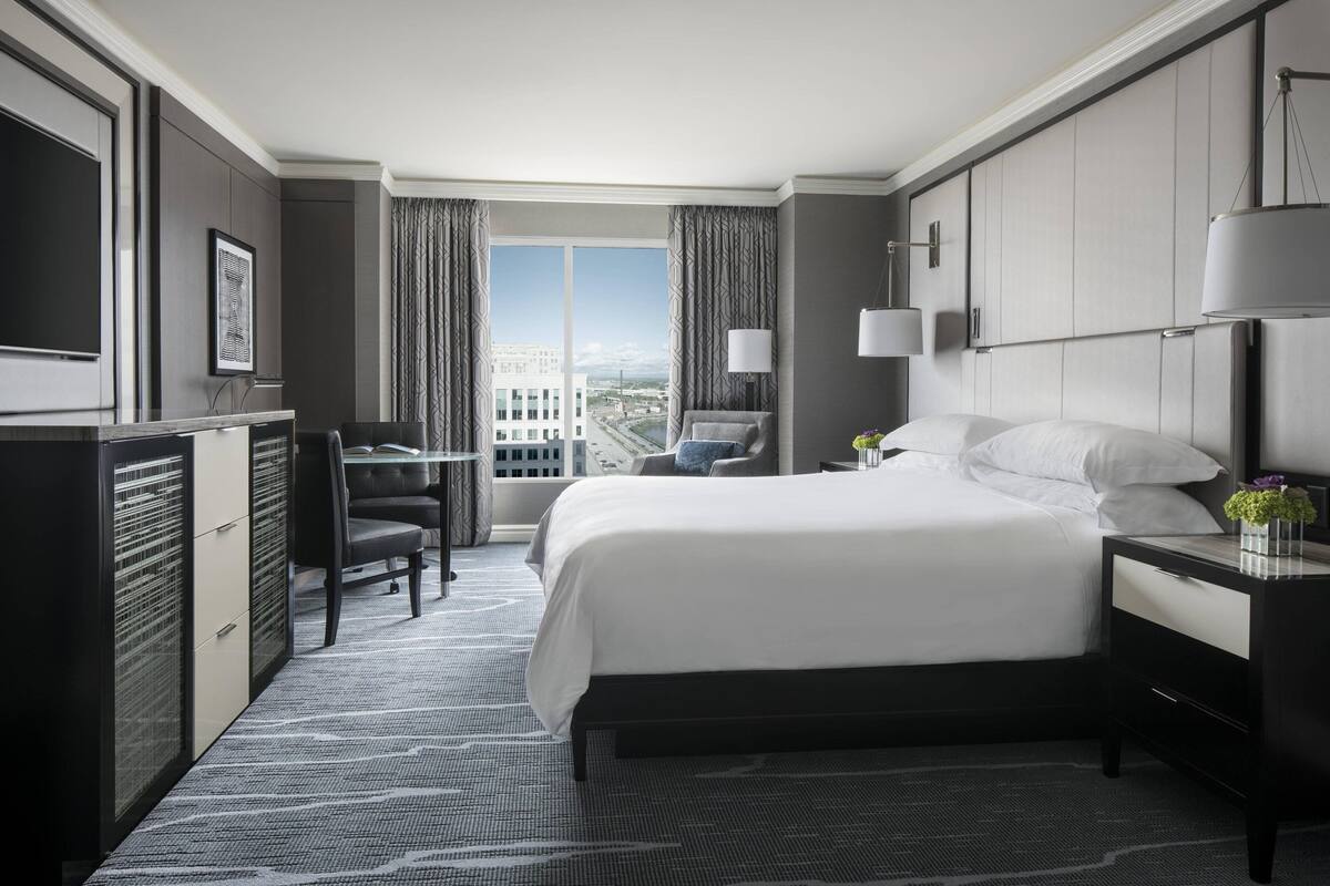 A hotel room with a large bed, a desk, a chair, a TV, and a view of the city.