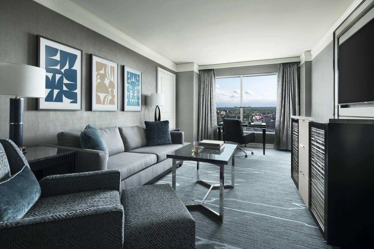 A modern hotel room with a sofa, armchairs, a glass coffee table, and a balcony with a city view.