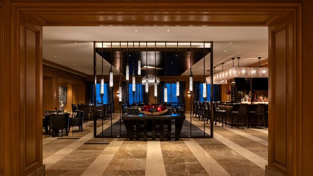 A spacious lobby with a central seating area, a bar on the right, and a view of the cityscape through large windows.