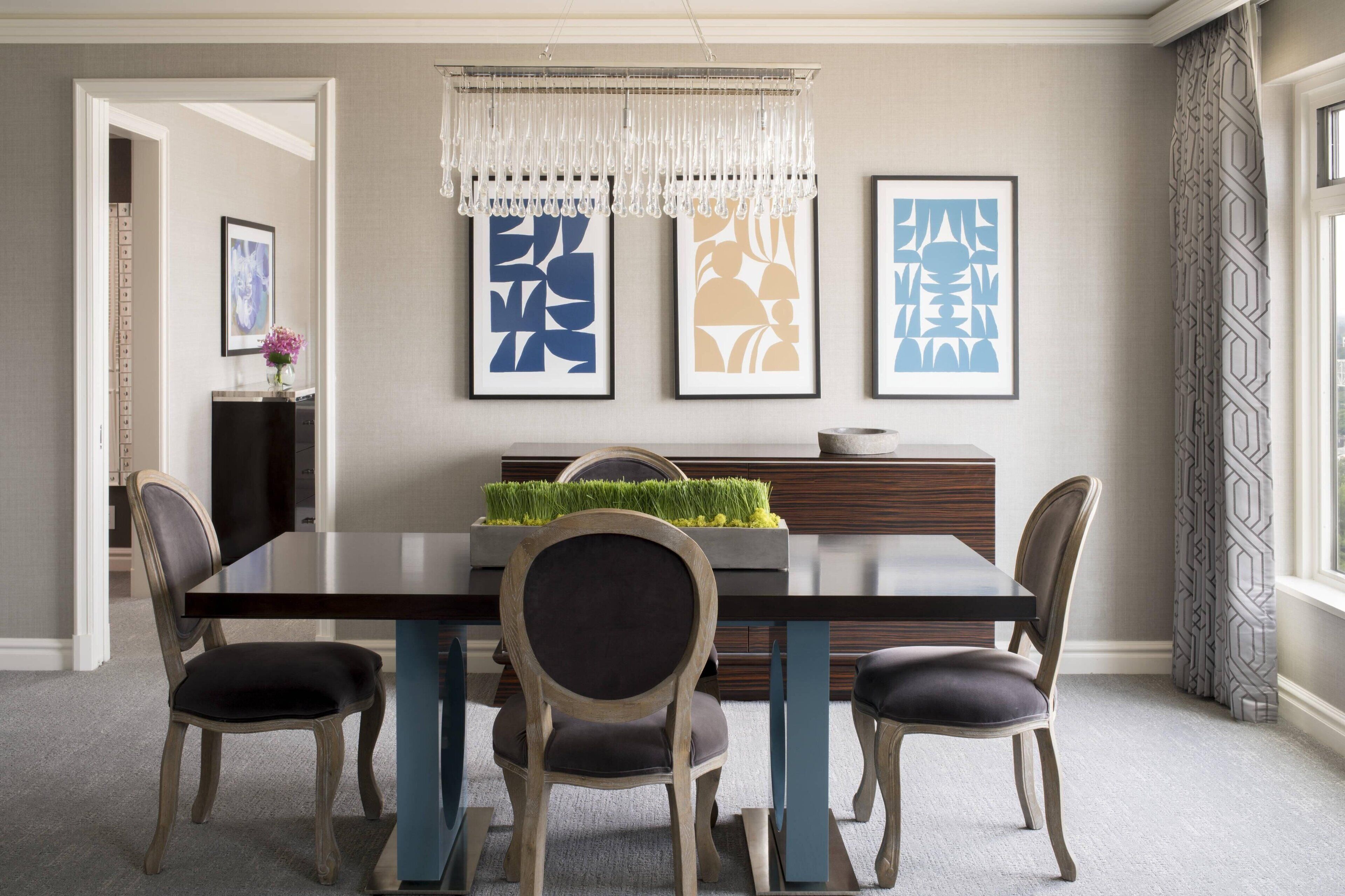 A dining area with a rectangular table, four chairs, a decorative hanging light, and framed artwork on the wall.