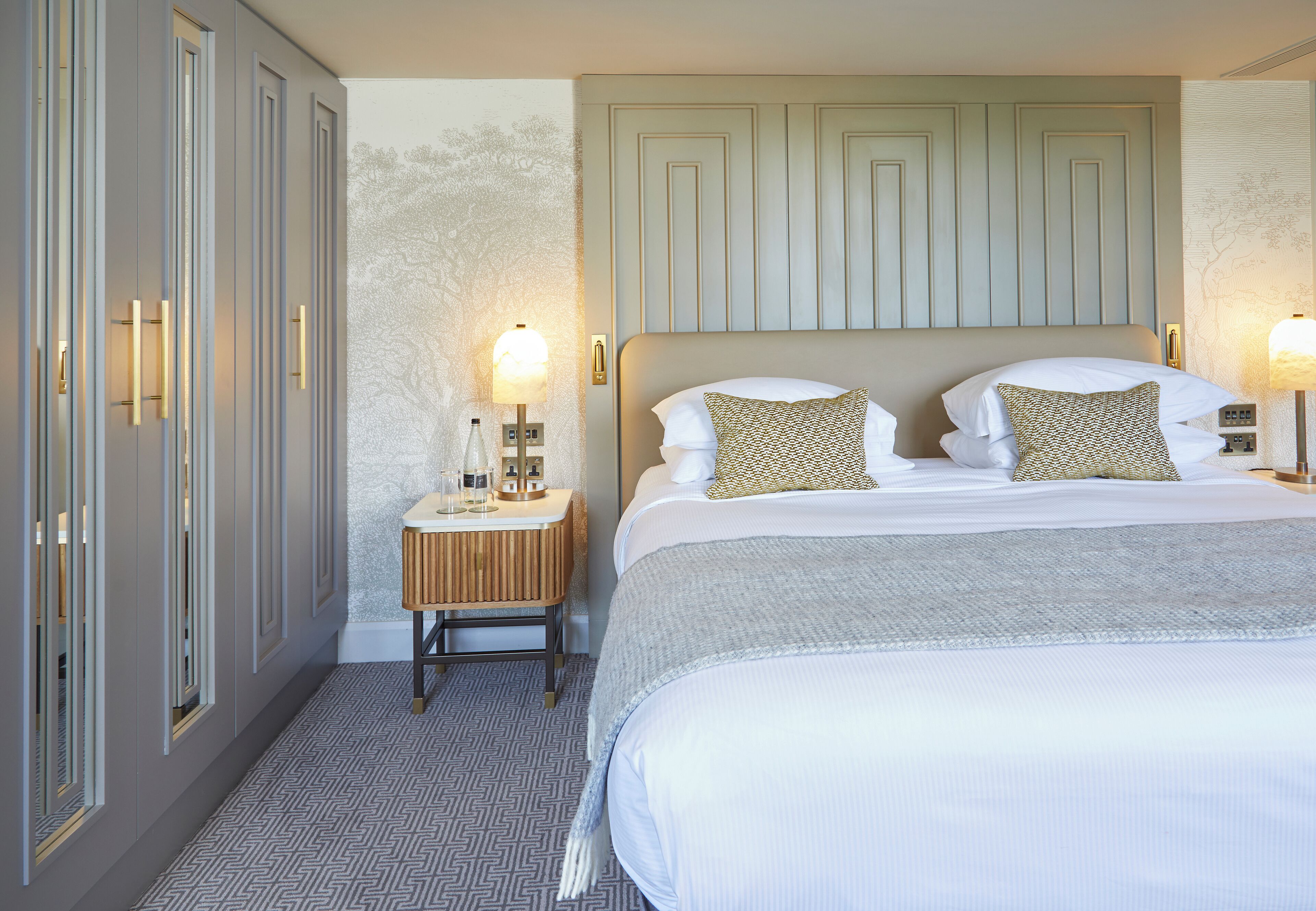 A hotel room with a large bed, a nightstand with a lamp, and a wardrobe with a mirror.