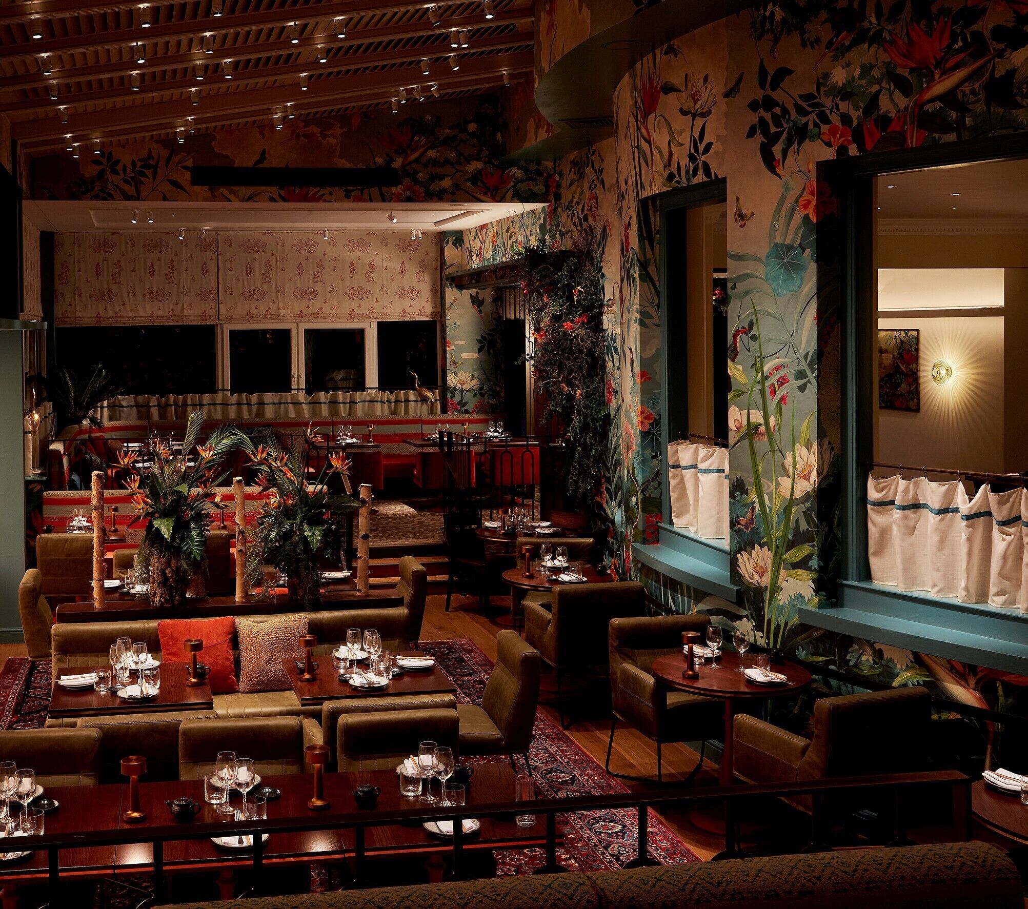 A restaurant with multiple dining areas, including booths and tables, decorated with floral patterns and plants.