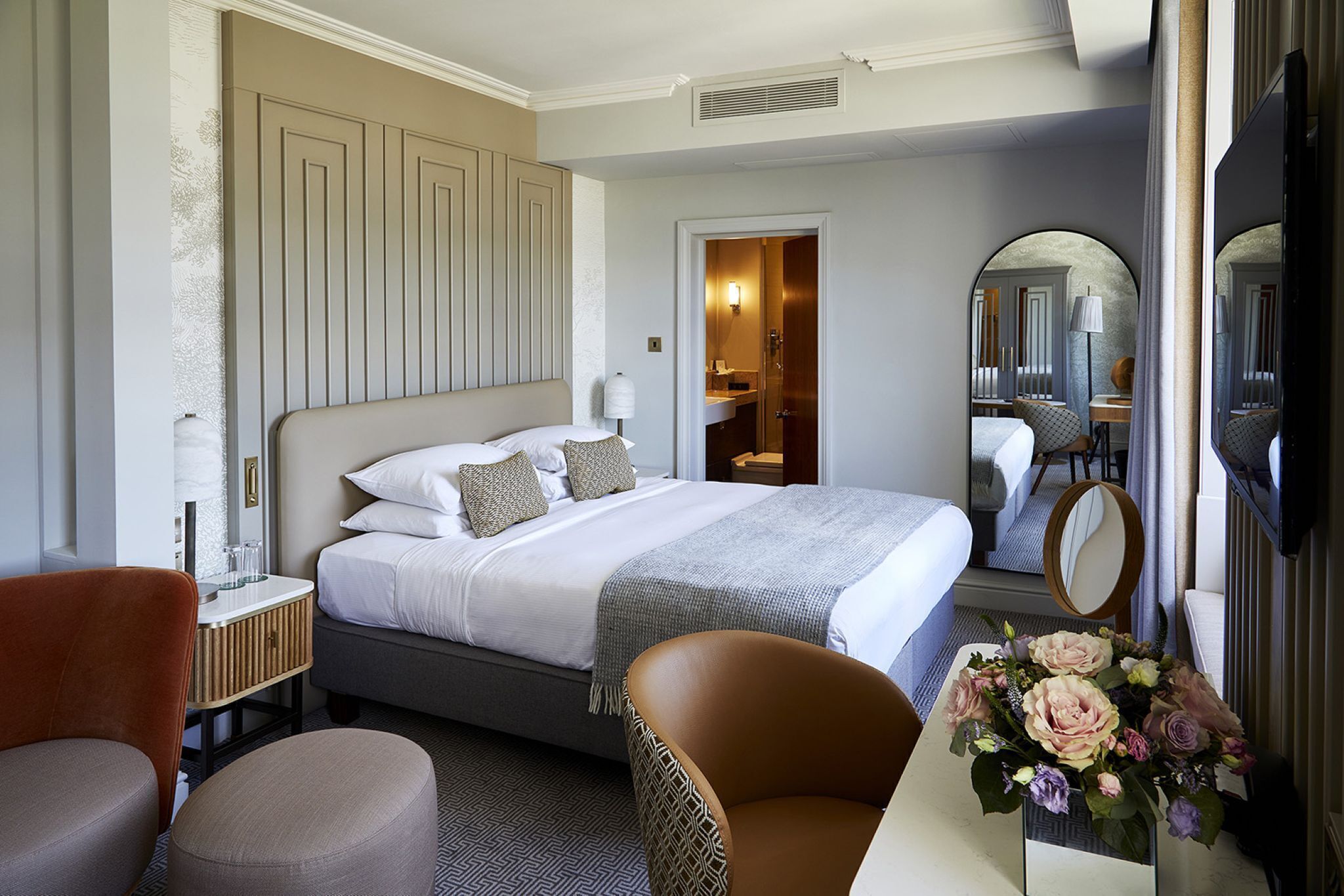 A hotel room with a bed, a desk, a chair, a mirror, and a vase with flowers.