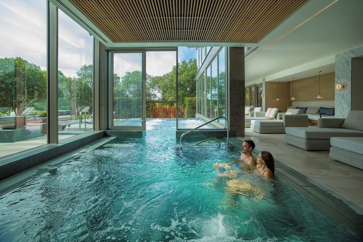 Ein moderner, überdachter Pool mit großen Glaswänden, einer Holzbalkendecke und einer Sitzecke mit Sofa und Fernseher.