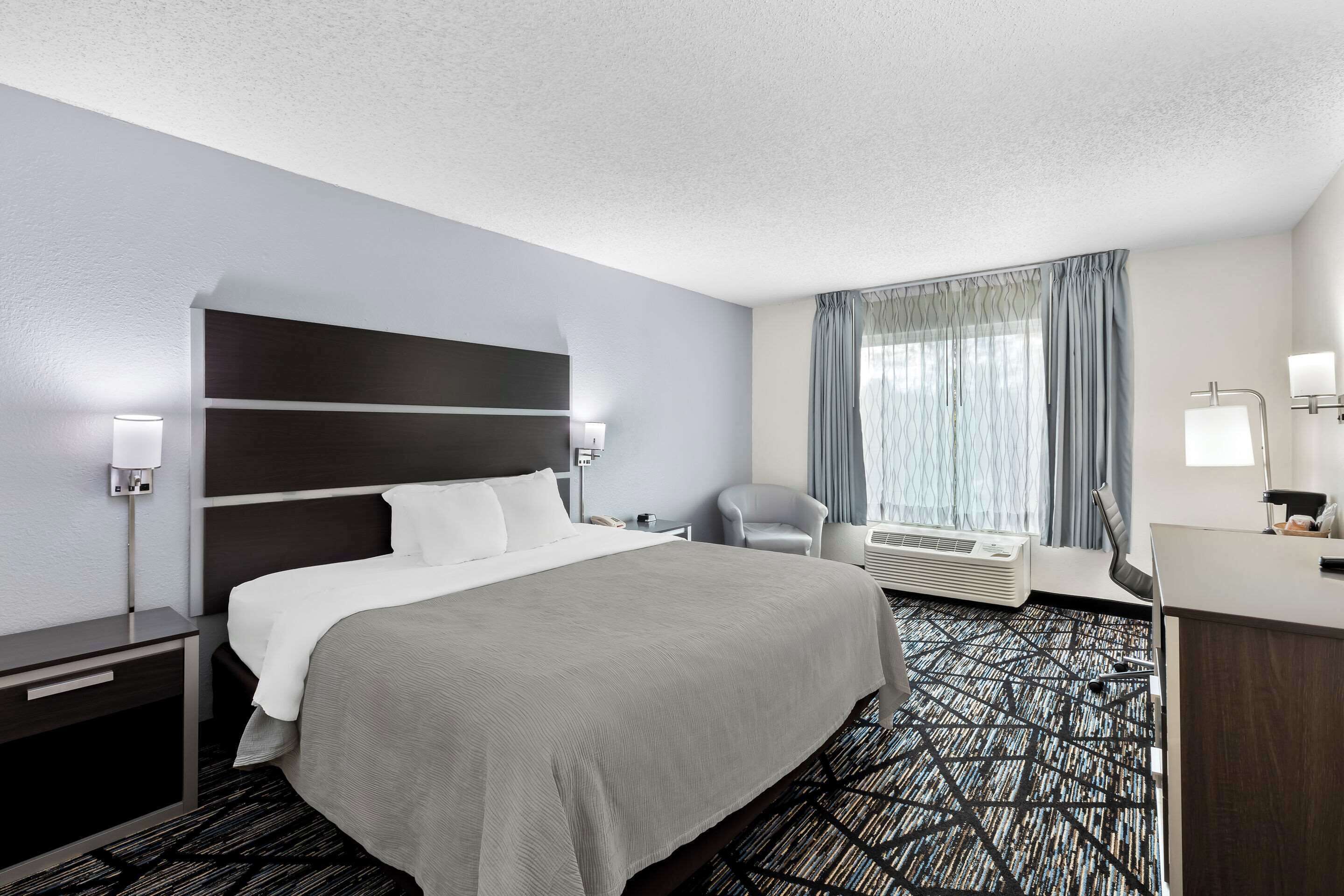A hotel room with a large bed, a desk, a chair, a window with curtains, and an air conditioning unit.