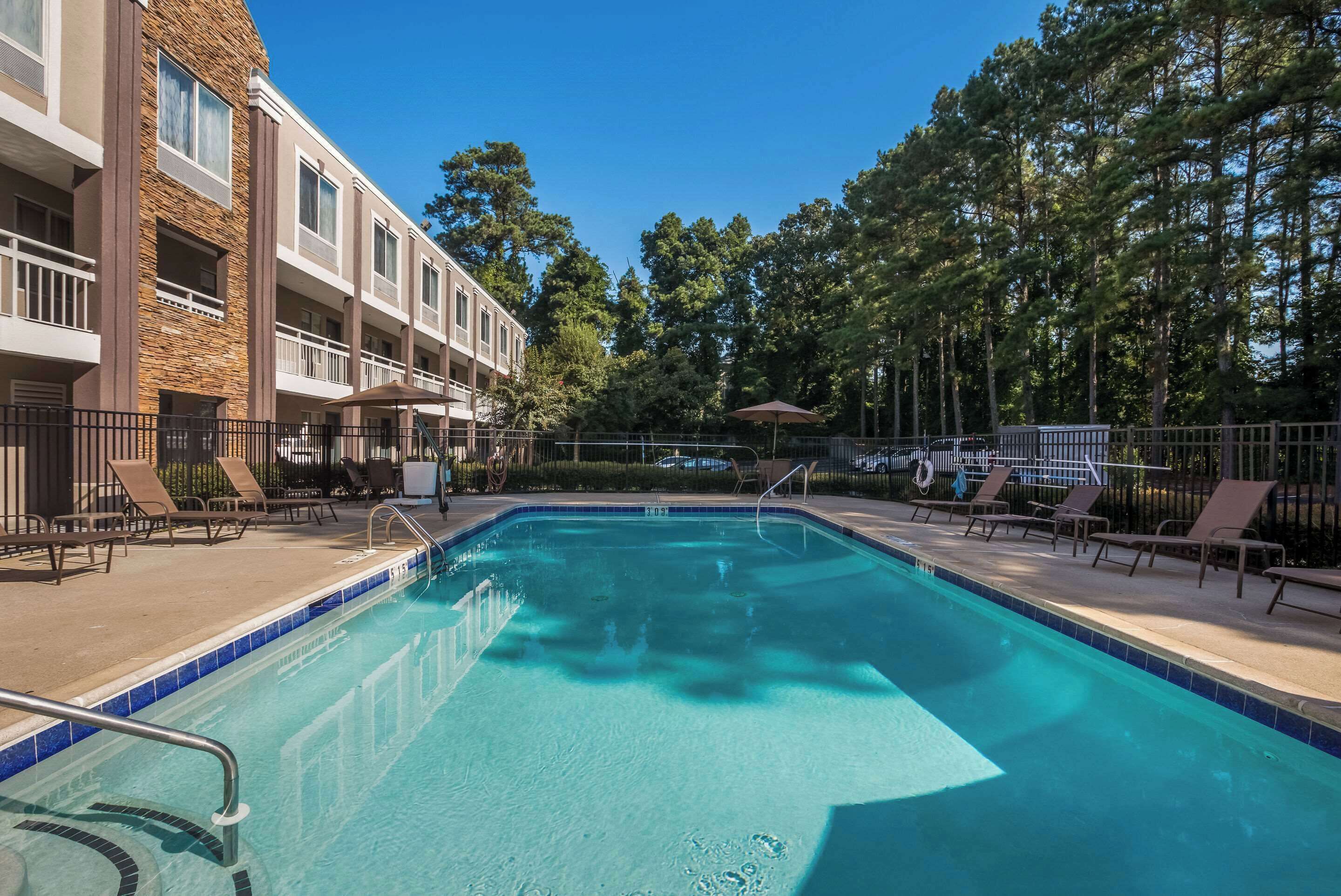 A swimming pool with lounge chairs and umbrellas, surrounded by a building with balconies and trees.