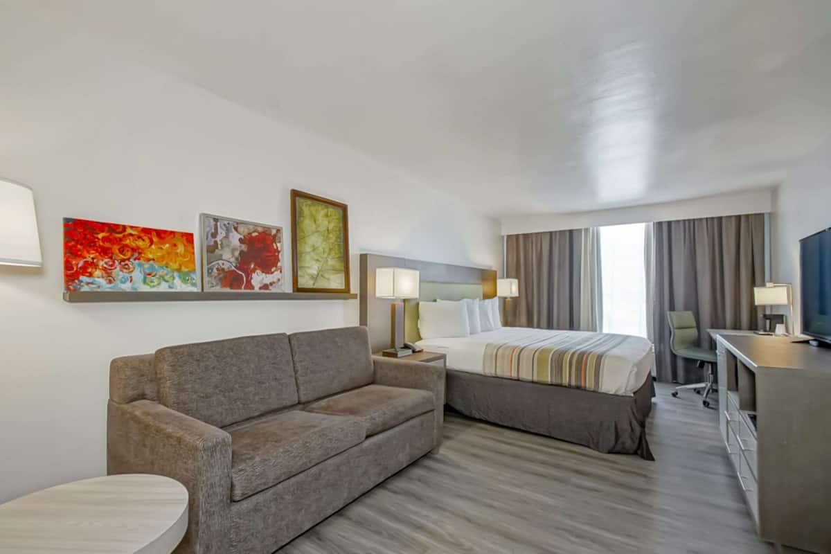 A hotel room with a sofa, a bed, a desk, and a TV. There are paintings on the wall and a window with curtains.