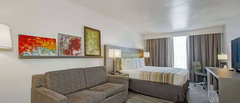 A hotel room with a sofa, a bed, a desk, and a TV. There are paintings on the wall and a window with curtains.