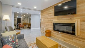 A modern living room with a flat-screen TV mounted on a wooden wall, a fireplace, a sofa with decorative pillows, and a dining area in the background. - Country Inn & Suites by Radisson, New Orleans I-10 East, LA (New Orleans)