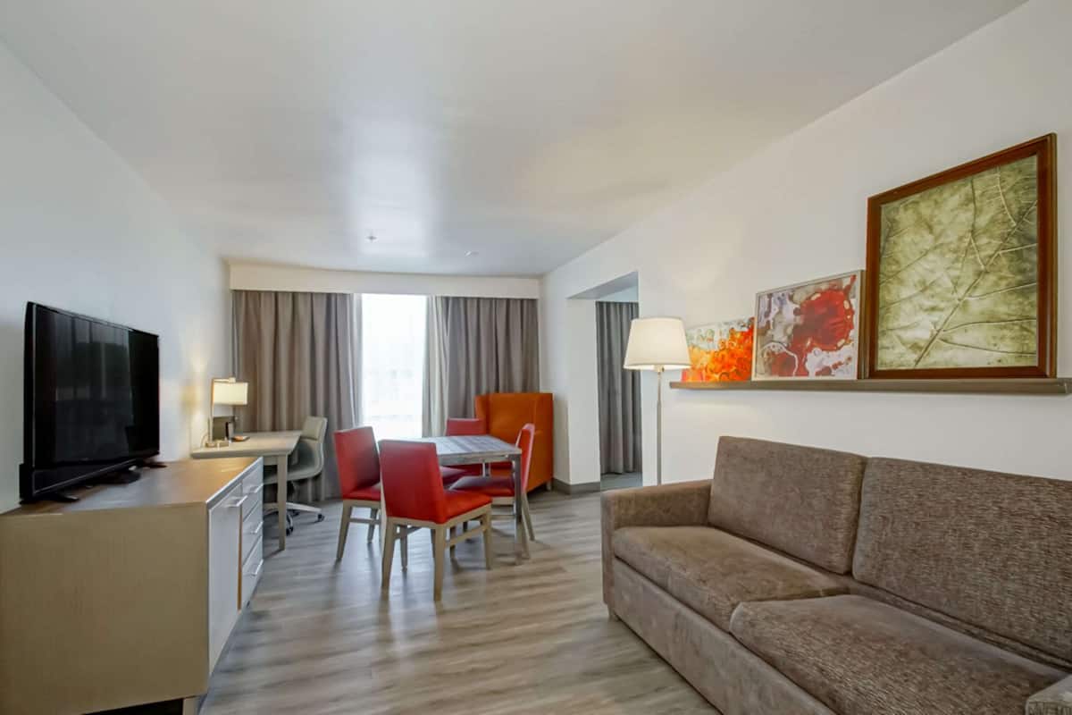 A hotel room with a flat-screen TV, a dining table with red chairs, a sofa, and artwork on the wall.