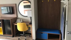 Ein Hotelzimmer mit Schreibtisch aus Holz, einem gelben Bürostuhl, einer blauen Bank, einem an der Wand befestigten Fernseher und einem Kleiderschrank mit Spiegel.