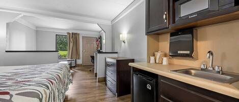 Habitación de hotel compacta con kitchenette, cama con ropa de cama estampada, un escritorio pequeño y vista al exterior a través de una ventana.
